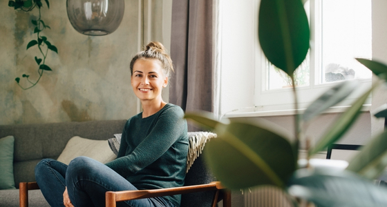 En kvinne sitter i en stol hjemme i en lys stue og smiler mot kameraet.