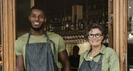To ansatte står i døråpningen til en kafé og smiler mot kameraet, iført forklær.