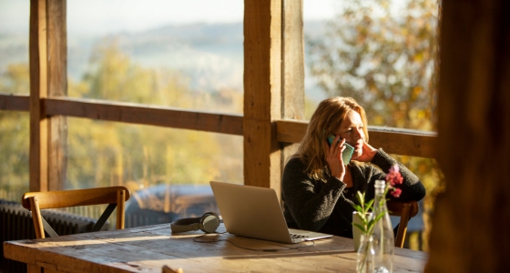 Kvinne sitter ved et bord med laptop og snakker i telefon i et solfylt rom med utsikt til naturen.