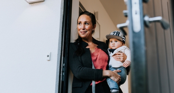 Kvinne st&aring;r i en d&oslash;r&aring;pning og holder et lite barn p&aring; armen.