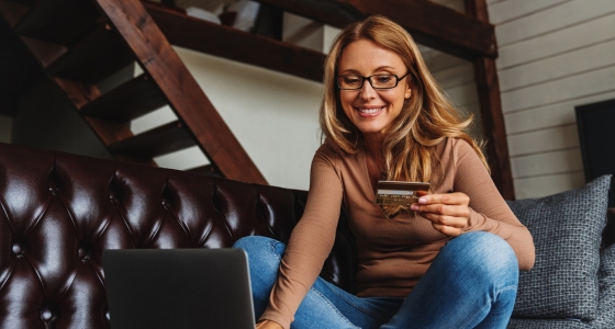 Kvinne sitter i sofaen og handler p&aring; nett med kredittkort og laptop.
