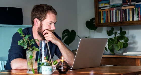 Mann sitter ved et bord hjemme og ser p&aring; laptop, omgitt av planter og b&oslash;ker.
