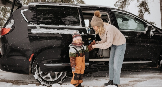 Mor hjelper barn ut av bilen i sn&oslash;v&aelig;r, begge kledd i varme vinterkl&aelig;r ved en parkert bil.