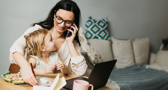 En kvinne sitter ved et bord hjemme med et barn, bruker laptop og snakker i telefonen.