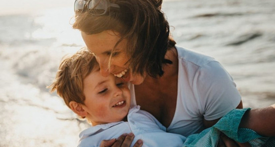 Mor som holder barnet sitt tett inntil seg ved sj&oslash;en, et varmt &oslash;yeblikk som symboliserer trygghet, omsorg og familieliv.