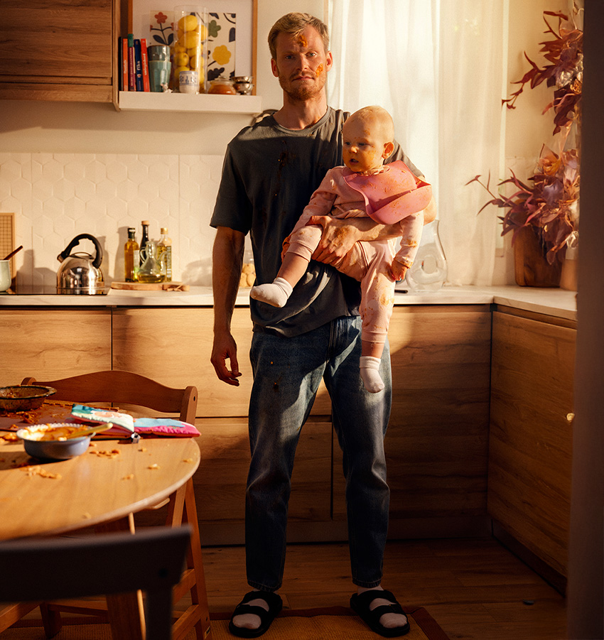 En pappaledig man står i ett stökigt kök med sin bebis i famnen, båda täckta av matspill.