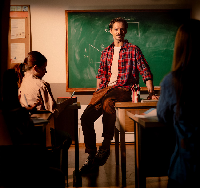 En vikarie sitter på katedern framför en griffeltavla och undervisar en klass.