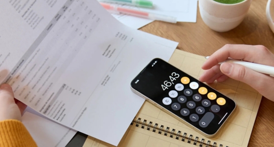 Person bruker kalkulator på mobil for å beregne utgifter og budsjett, med regninger og notater på bordet.