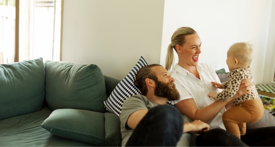 Et par sitter i sofaen og leker med babyen sin i en lys stue.