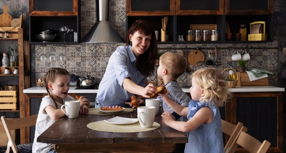 Mor sitter ved kj&oslash;kkenbordet sammen med tre barn og serverer frokost i et hjemmekj&oslash;kken.