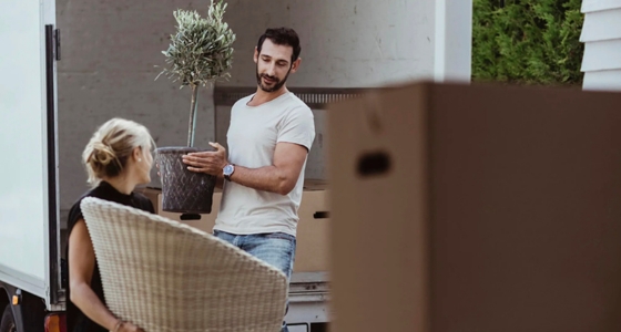 Personer som flytter inn i ny bolig, der en mann bærer en potteplante fra en flyttebil mens en annen person holder en stol.