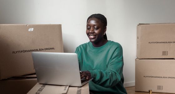 En kvinne sitter p&aring; gulvet blant flytteesker og bruker en laptop i en tom bolig.