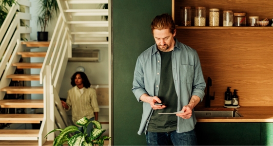 Mann som st&aring;r hjemme og ser p&aring; mobiltelefon og et dokument, mens en annen person er synlig i bakgrunnen av boligen.