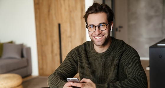 Smilende mann med briller som sitter hjemme og bruker mobiltelefon, et bilde som symboliserer enkel og digital bankhverdag.