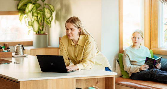Mor jobber hjemme p&aring; laptop ved kj&oslash;kkenbenken mens barn sitter og leser i bakgrunnen